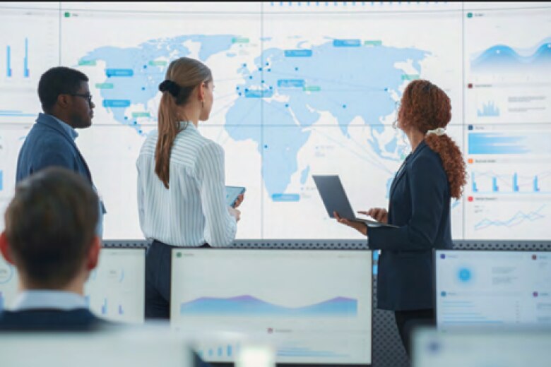 Three supply chain executives analyze supply chain data on a large map with several charts of data
