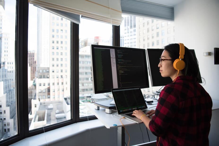 IT developer with orange headphones looking out their office window, while working on multiple monitors