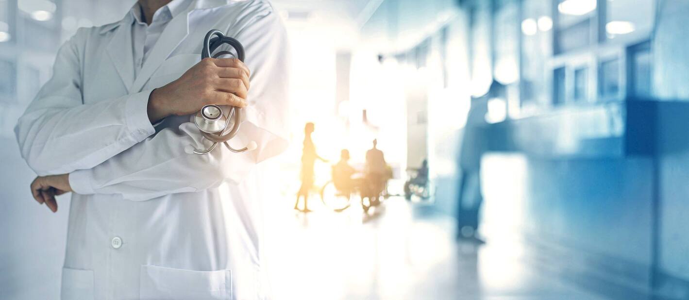 Doctor with arms crossed holding stethoscope in foreground of hospital hallway