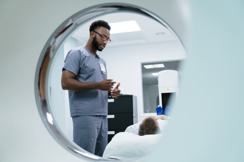 Male nurse talking with patient lying in examination room seen through MRI Scanner