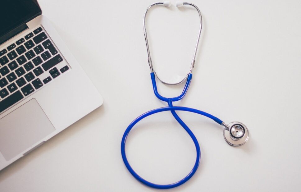 blue stethoscope lies on a white table surface next to an open laptop