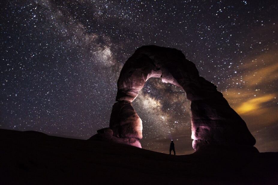 Person standing under a massive rock formation underneath an expansive sky full of stars