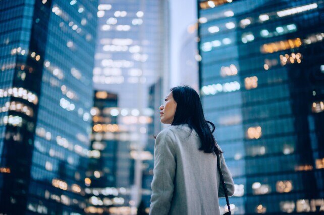 side profile of confident and professional young Asian businesswoman looking up while standing against contemporary corporate skyscrapers