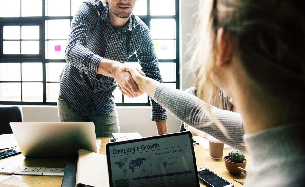 Two coworkers shaking hands over their laptops from either side of a table