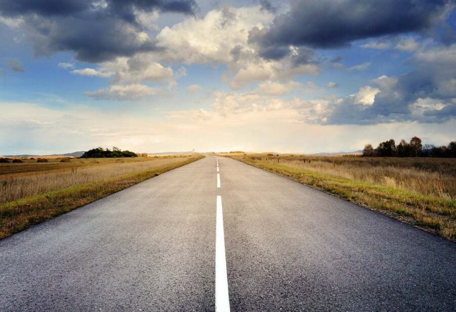 wide open road extending to the horizon under a cloudy blue sky with open fields on either side