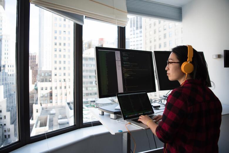 woman working behind multiple computer screens while wearing headphones and looking out over city