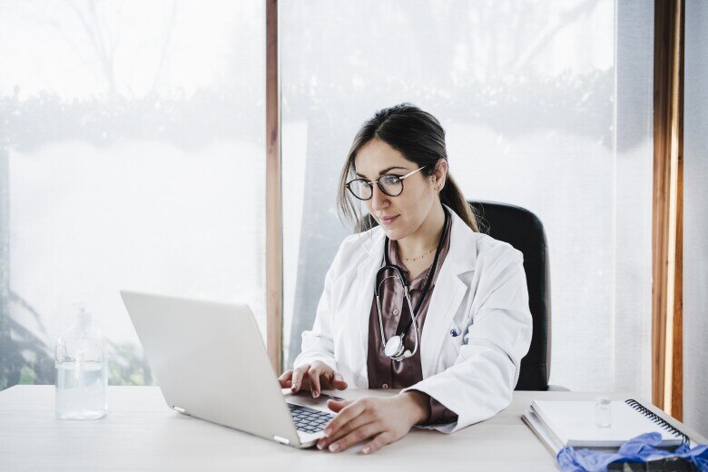 Female medical doctor using laptop while sitting against window in hospital