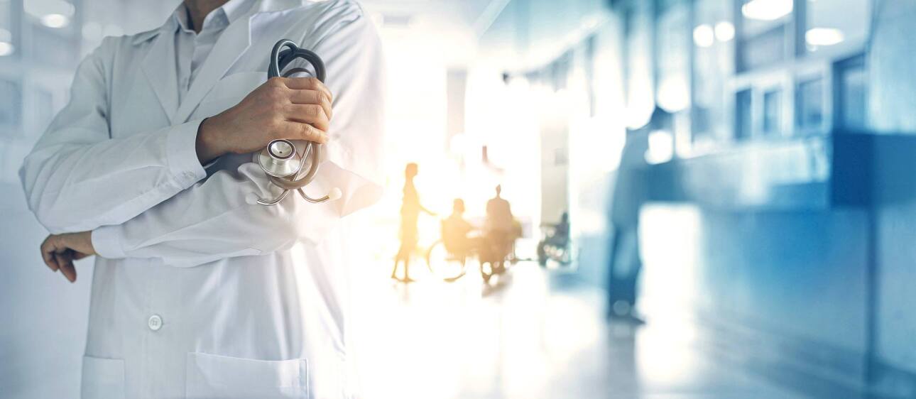 Doctor with arms crossed holding stethoscope in foreground of hospital hallway