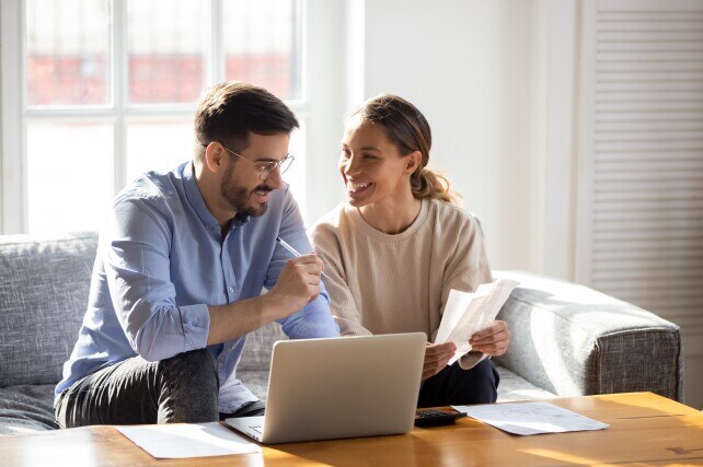 Happy young man and woman sitting on a couch looking at their finances on a laptop and paper