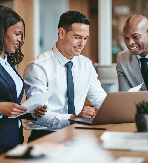 Smiling group of diverse businesspeople going over paperwork together and working on a laptop at a table in an office