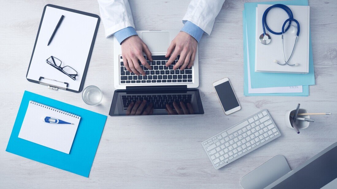 aerial view of a medical doctor typing on the keyboard of a laptop with notes nearby on desk