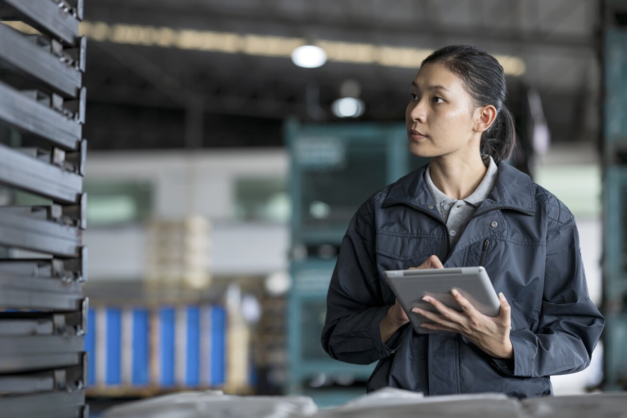 An Asian female engineer holding a tablet computer and standing near a car chassis on a pallet to manage and control for supply to production line assembly.